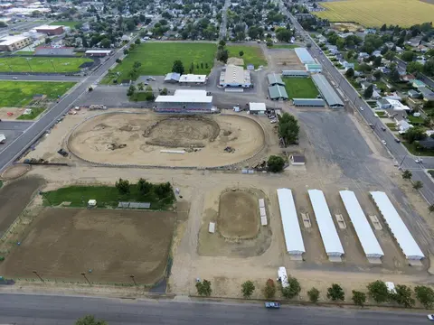An aerial view shows an outdoor fairgrounds complex with a racetrack, stables, and surrounding town.