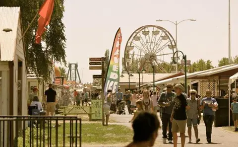 People walking along a fairground midway with a Ferris wheel in the background.