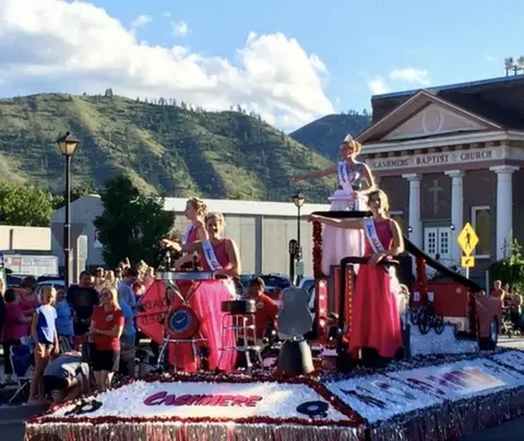 A decorated parade float carries women in pink dresses past a crowd.