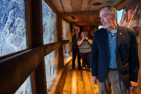 A man looks ahead while walking through a wooden indoor exhibit space with other visitors in the background.