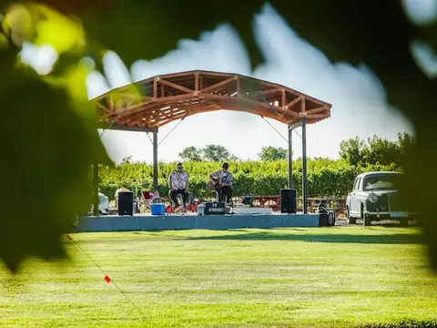 Musicians perform on an outdoor stage in a grassy field next to a vintage car.