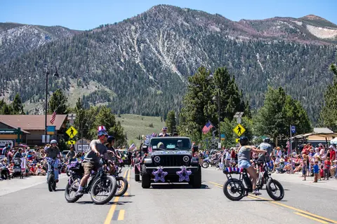 A parade featuring bicycles and a jeep with patriotic decorations moves down a street with a large mountain in the background.