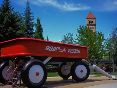 A large red toy wagon sits outdoors near trees and a tower.