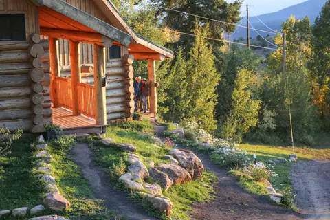 A log cabin sits in a landscape with two people standing on its porch.