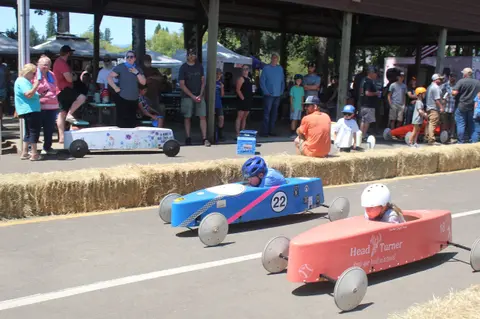 Children race homemade soapbox cars down a track lined with hay bales while spectators watch.
