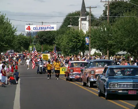 A parade on a town street with classic cars, people walking, and flags.