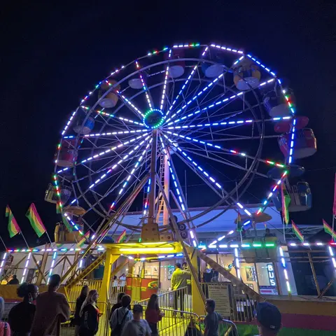 A brightly lit Ferris wheel at night with people gathered around its entrance.