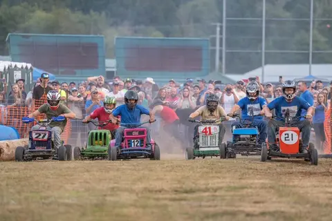 Several racers compete in a lawn mower race before a crowd of spectators.