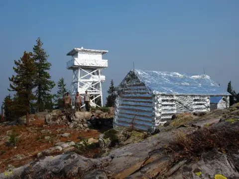 Three people stand near a fire lookout tower and a cabin wrapped in reflective foil.