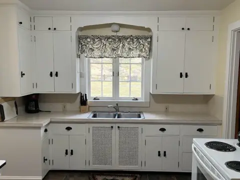 A photograph of a kitchen interior featuring white cabinets and a window above the sink.