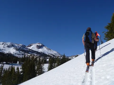 Two skiers climb a snow-covered slope toward a mountain peak.