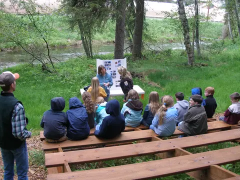 An outdoor presentation taking place by a river with children seated on wooden benches.