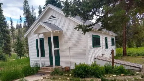 A small white building with green trim sits in a grassy, wooded area.