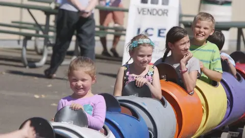 Children riding a colorful barrel train at an outdoor event.
