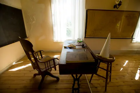 A historic classroom interior featuring a wooden desk, chair, and a dunce cap on a stool.