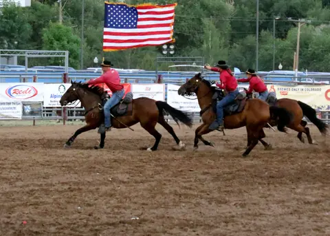 Three riders on horses perform in an arena carrying an American flag.