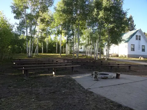 An outdoor amphitheater with wooden benches surrounded by tall trees and a building nearby.