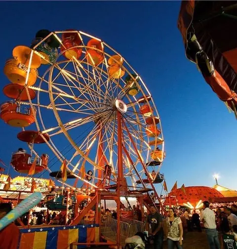 A brightly lit Ferris wheel stands at a carnival at dusk.