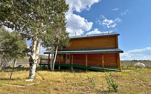 A two-story log building with a covered porch and ramp stands next to trees under a partly cloudy sky.