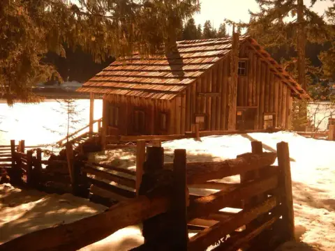 A rustic wooden cabin sits in a snowy landscape behind a wooden fence.