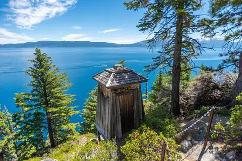 A small wooden outhouse overlooks a large blue lake surrounded by mountains and trees.