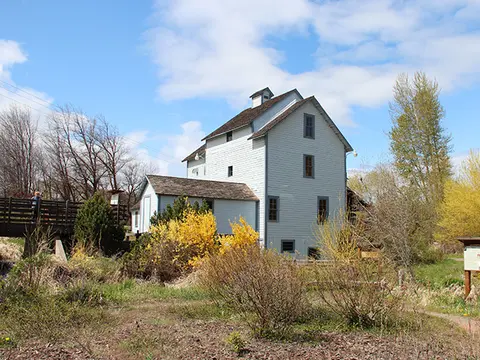 An exterior view of a historic wooden mill building surrounded by trees and bushes under a partly cloudy sky.