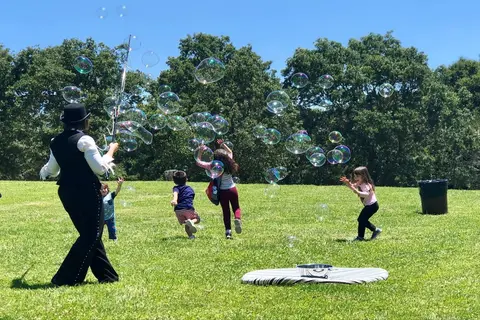 A person blowing giant bubbles outdoors in a grassy field with children chasing them.