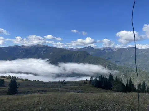 A scenic mountain landscape with low-hanging clouds over forested slopes under a blue sky.