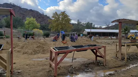 People working in a dirt lot outdoors with wooden mining tables and shelters.