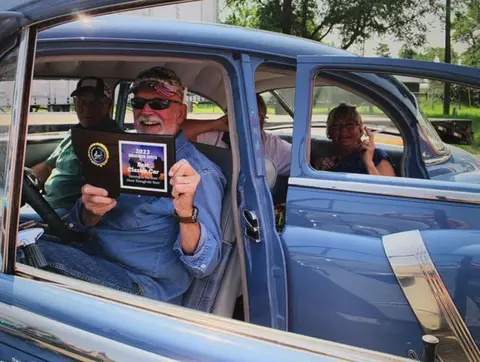 People sitting inside a classic blue car holding a plaque.