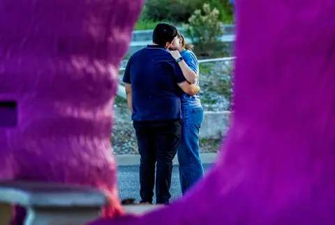 A couple embraces and kisses outdoors framed by bright purple foreground objects.
