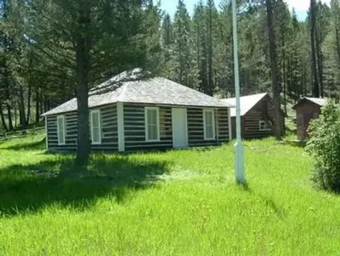 A log building stands in a grassy clearing surrounded by trees.