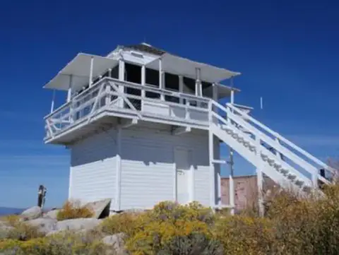 A white wooden fire lookout tower stands on a rocky hillside against a clear blue sky.