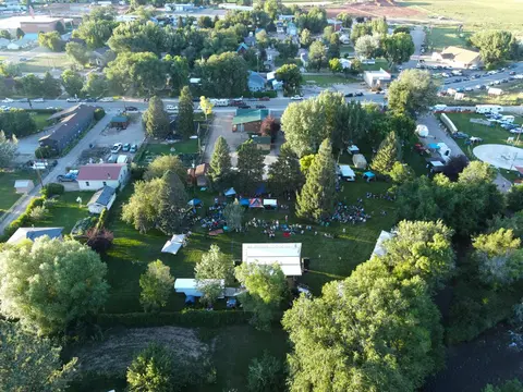 An aerial view shows an outdoor gathering with a stage and tents set up on a grassy lawn among trees.