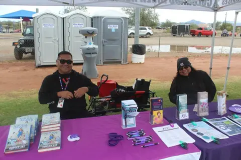 Two people sit behind a purple table with books and pamphlets at an outdoor event.