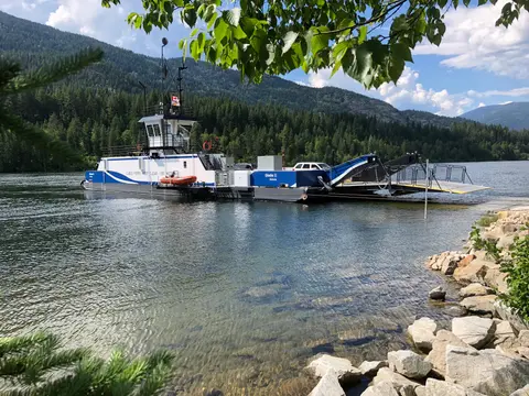 A cable ferry floats on a calm river with forested mountains in the background.