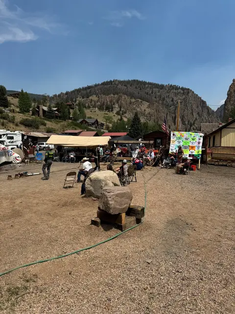 People gather in a gravel area surrounded by buildings and mountains.