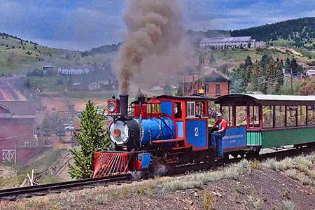 A small colorful steam locomotive pulls passenger cars along tracks through a hilly landscape.