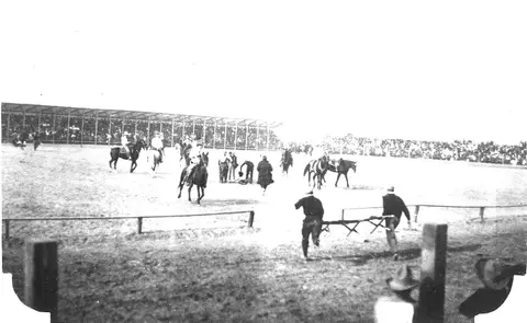 A historic black and white photograph showing people and horses in an outdoor arena.