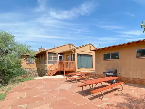 An exterior view of a rustic building with a stone patio, picnic tables, and a grill.