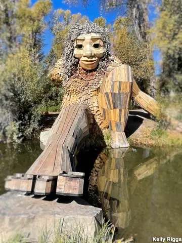 A large wooden troll sculpture sits near a body of water with trees in the background.