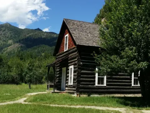 A log cabin stands in a grassy clearing with mountains in the background.