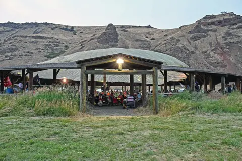 An outdoor circular structure with people gathered underneath, set against a barren hill landscape.