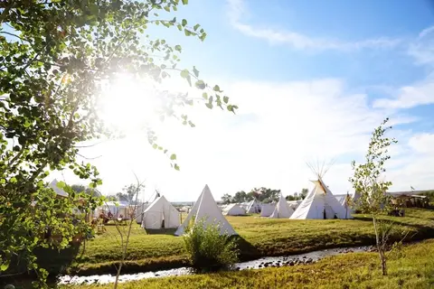 A campsite with tipis and tents set up in a grassy field near a stream under a bright sun.