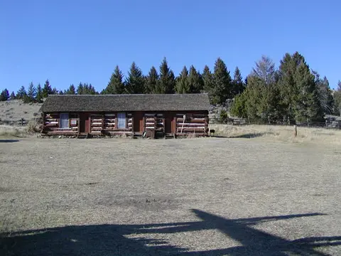 A long single-story log cabin sits in a dry grassy field backed by evergreen trees.