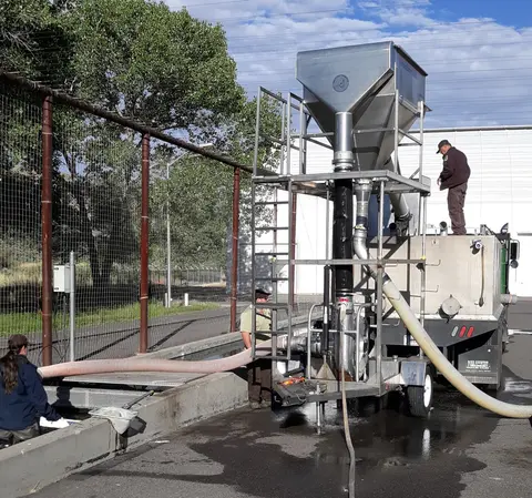 Workers operate a specialized fish transfer truck and equipment outside near a fence.
