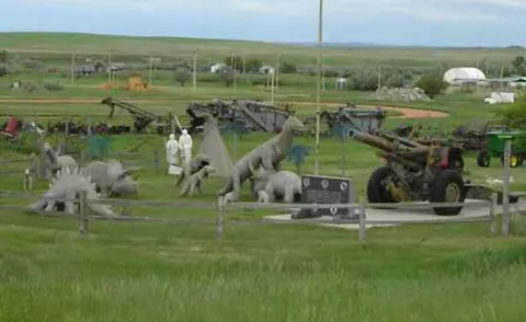 An outdoor display featuring dinosaur statues, old artillery, and farm machinery in a grassy landscape.
