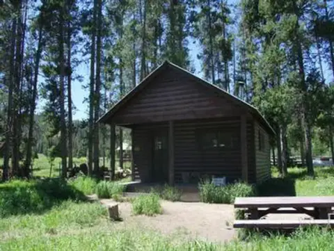 A wooden cabin sits surrounded by tall trees and a grassy clearing.