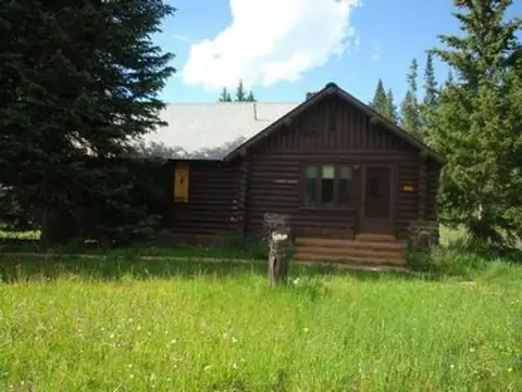 A rustic log cabin sits in a grassy clearing surrounded by trees.