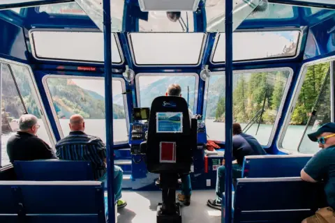 Interior view of passengers seated on a tour boat with large windows looking out at water and mountains.